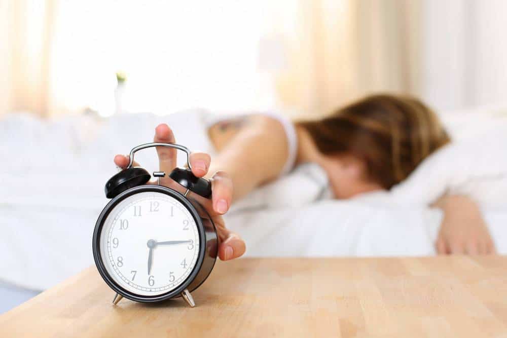 A woman pushing her alarm clock while getting out of bed.