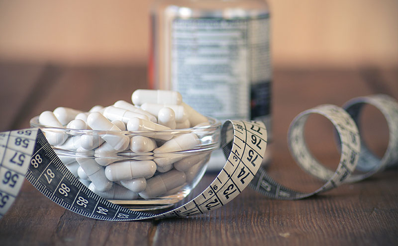 Tape measure with white capsules in a glass bowl in front of a supplement box, on dark wooden table